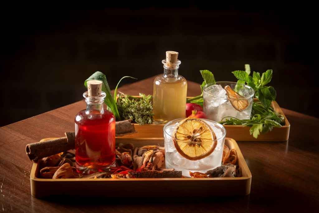 A wooden tray displaying various drinks, including two glasses with ice and garnishes, and three glass bottles filled with colorful liquids, surrounded by herbs and spices.